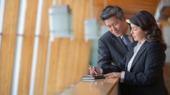 Mature Caucasian businesswoman and Chinese Asian businessman standing on office balcony taking notes 