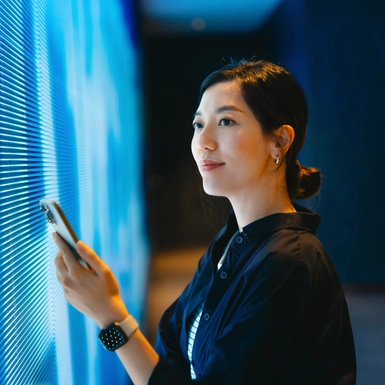 Confident young Asian woman using smartphone against blue coloured illuminated LED digital display screen in the dark