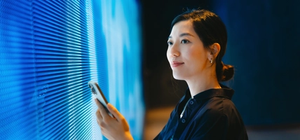 Confident young Asian woman using smartphone against blue coloured illuminated LED digital display screen in the dark