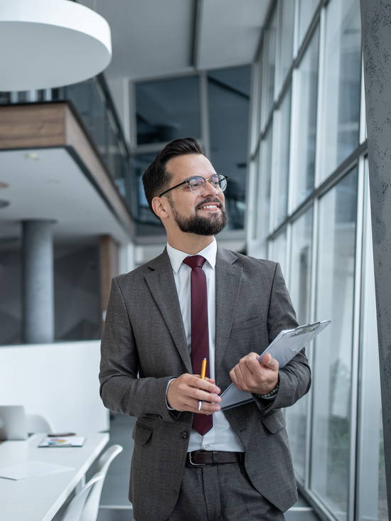 Smiling businessman holding clipboard and pen, looking away in modern office