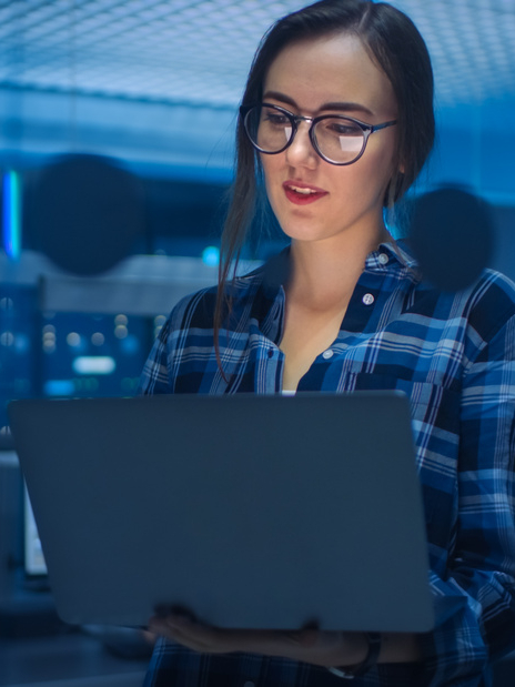 Portrait of a Smart Seductive Young Woman Wearing Glasses Holds Laptop. In the Background Technical Department Office with Specialists Working and Functional Data Server Racks