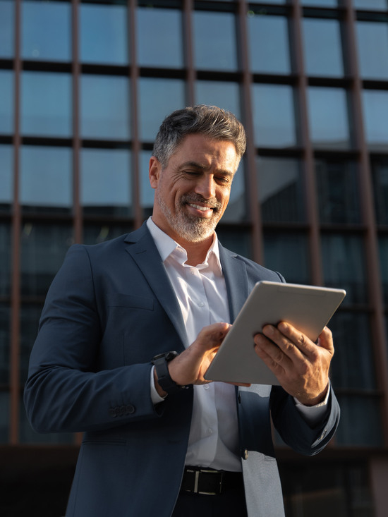 Focused successful mature Indian or Latin entrepreneur businessman holding digital pc tablet standing outdoor at business office building. Hispanic smiling man in suit working using touchpad computer
