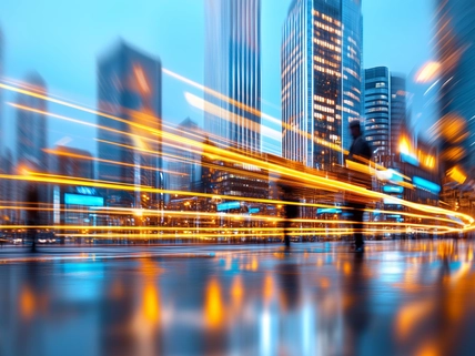 Blurred business professionals crossing rainy city street, vibrant light trails illuminating nighttime urban landscape with skyscrapers towering overhead