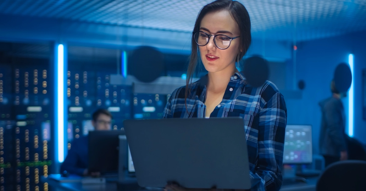 Portrait of a Smart Seductive Young Woman Wearing Glasses Holds Laptop. In the Background Technical Department Office with Specialists Working and Functional Data Server Racks