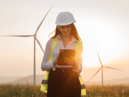 Beautiful caucasian woman in white helmet working with digital tablet at renewable energy farm. Female inspector controlling functioning of wind turbines outdoors.