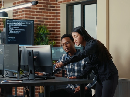 Two software developers holding laptop with coding interface walking towards desk and sitting down