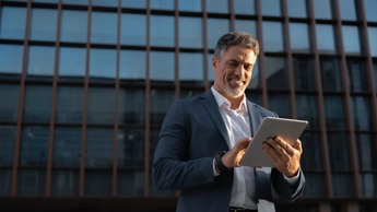 Focused successful mature Indian or Latin entrepreneur businessman holding digital pc tablet standing outdoor at business office building. Hispanic smiling man in suit working using touchpad computer