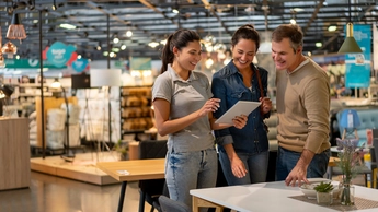 Cheerful sales woman showing a design on tablet to mid adult couple looking for furniture at a home store