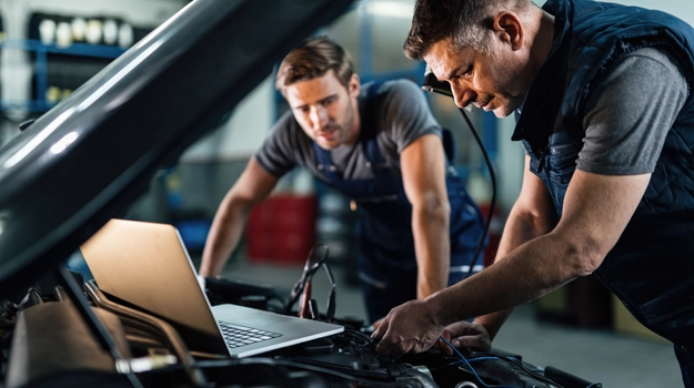 two men working on a car with a laptop to assist
