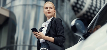Businesswoman wearing black suit using smartphone, leaning on electric car recharge battery at charging station in city