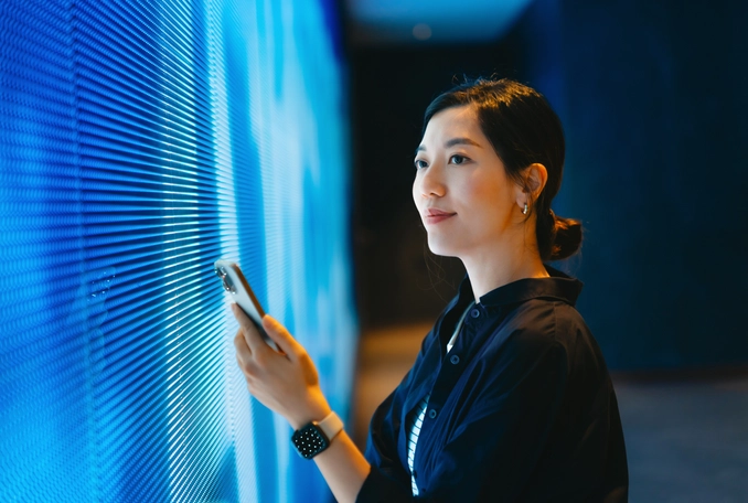 Confident young Asian woman using smartphone against blue coloured illuminated LED digital display screen in the dark