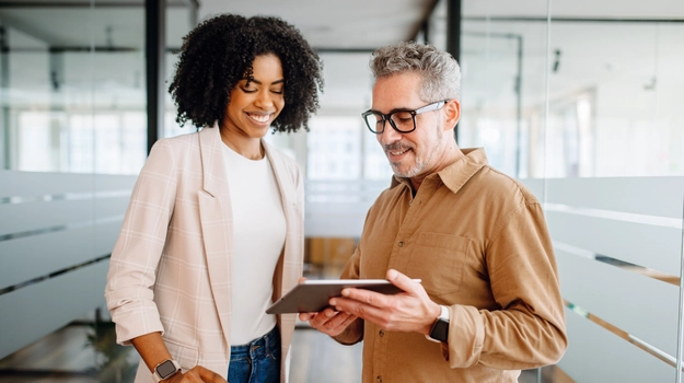 A businesswoman and a senior male colleague collaboratively review content on a tablet in a bright office space