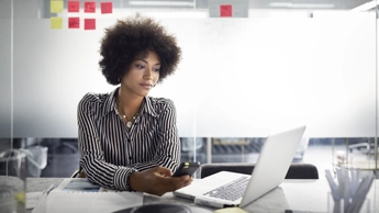 Businesswoman looking at laptop computer while holding smart phone in office