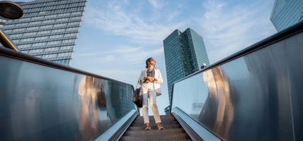 Woman on escalator going down with buildings and sky behind her