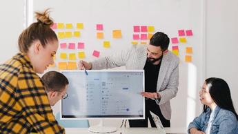 Man in office pointing at computer screen while explaining to colleagues